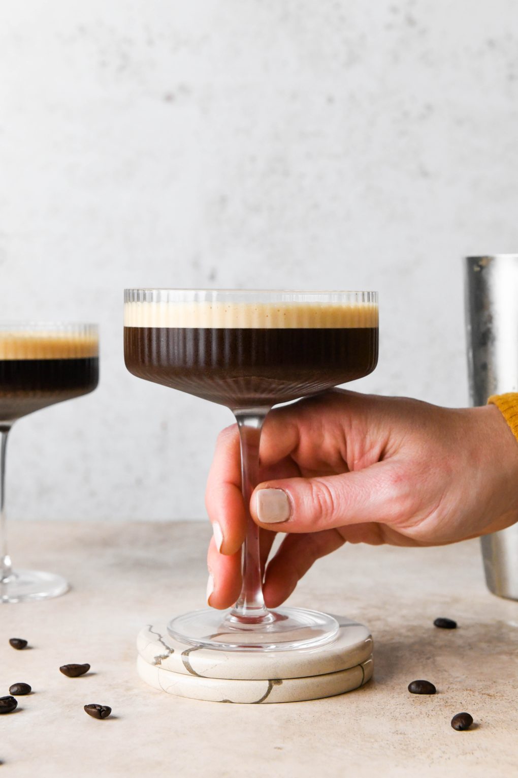 Hand reaching into the frame to grasp the stem of a coupe glass of espresso martini.