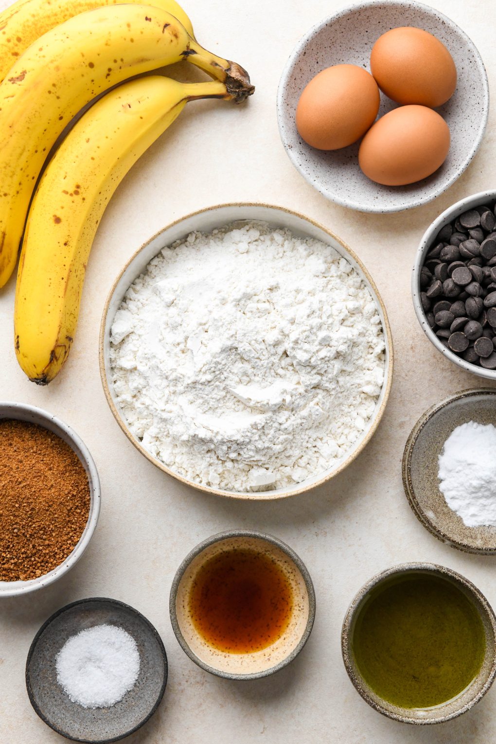 Ingredients for easy gluten free banana bread in various bowls on a cream colored surface.