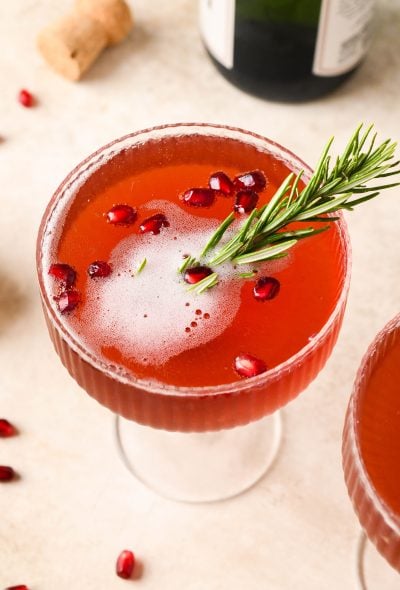 Overhead shot of festive pomegranate champagne cocktail garnished with pomegranate seeds and a sprig of rosemary. On a light brown background next to another cocktail and scattered pomegranate seeds.