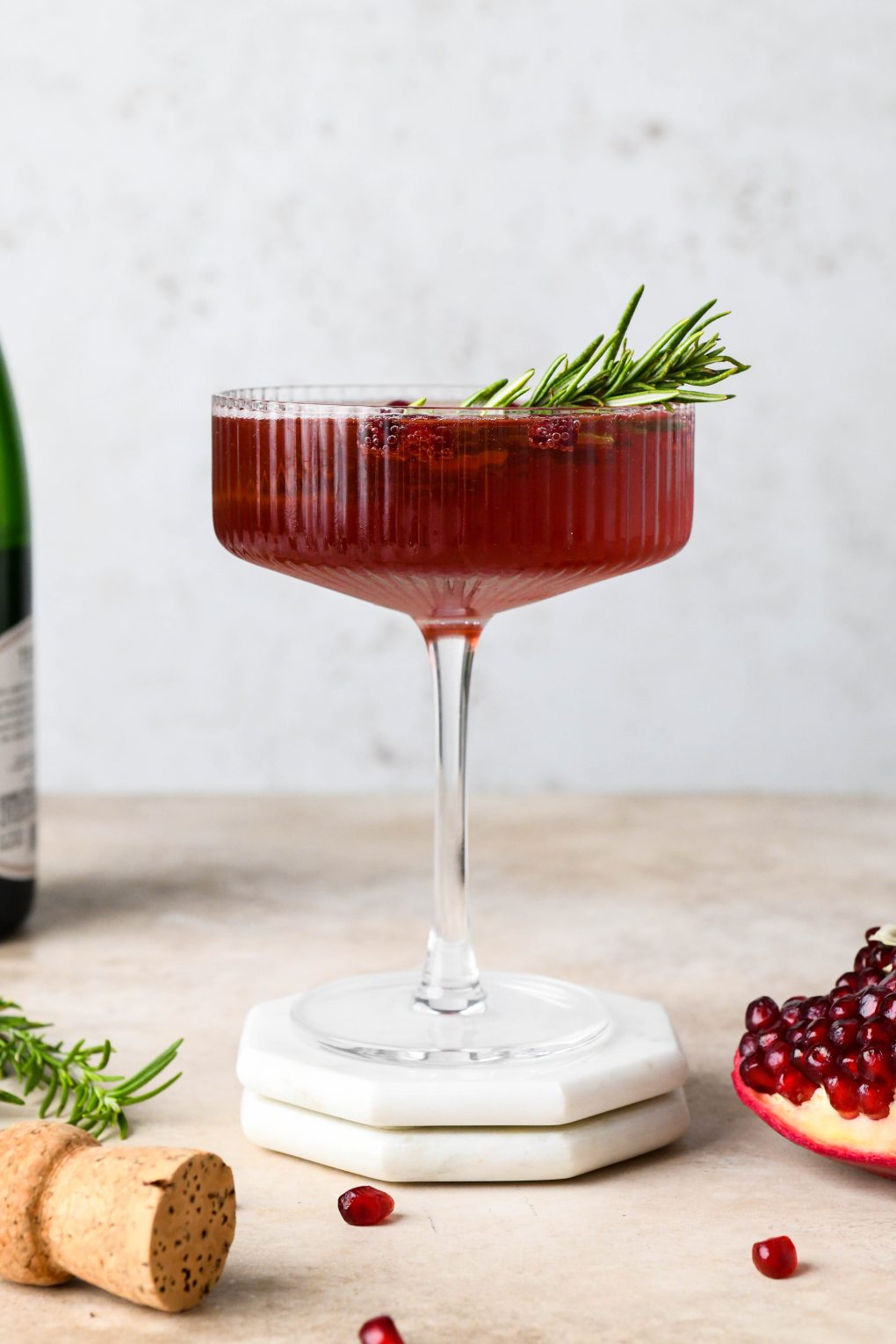 Straight on shot of a bright red bubbly festive pomegranate champagne cocktail garnished with a sprig of rosemary and sitting next to a piece of pomegranate fruit and a champagne cork.