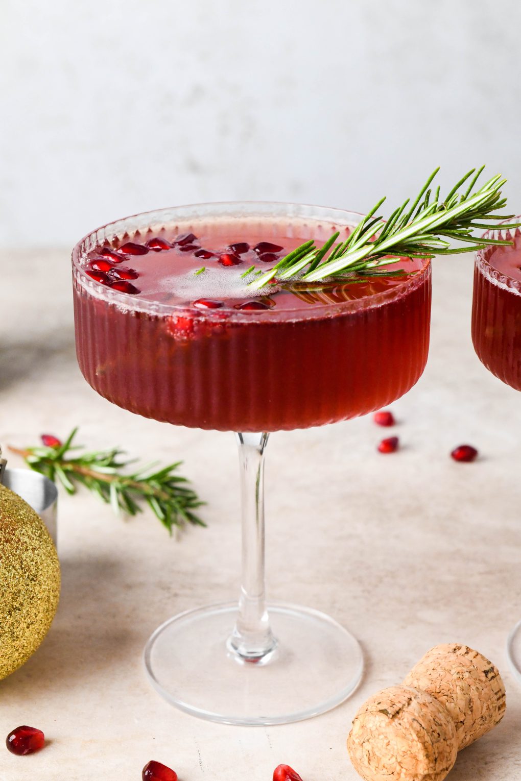 Straight on shot of a bright red bubbly festive pomegranate champagne cocktail garnished with a sprig of rosemary and sitting next to a piece of pomegranate fruit and a champagne cork.