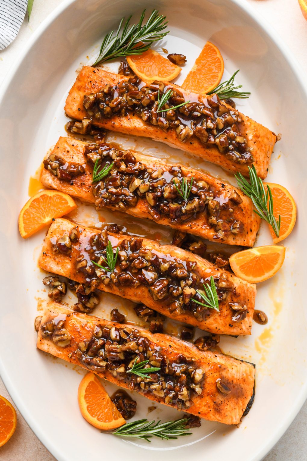 Oven roasted maple pecan glazed salmon in an oval cream colored baking dish. Garnished with orange slices and fresh rosemary. 