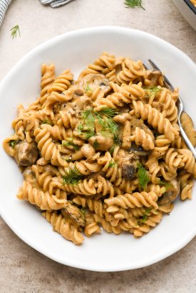 A white ceramic plate filled with creamy vegan mushroom stroganoff made with fusilli pasta and topped with fresh dill.