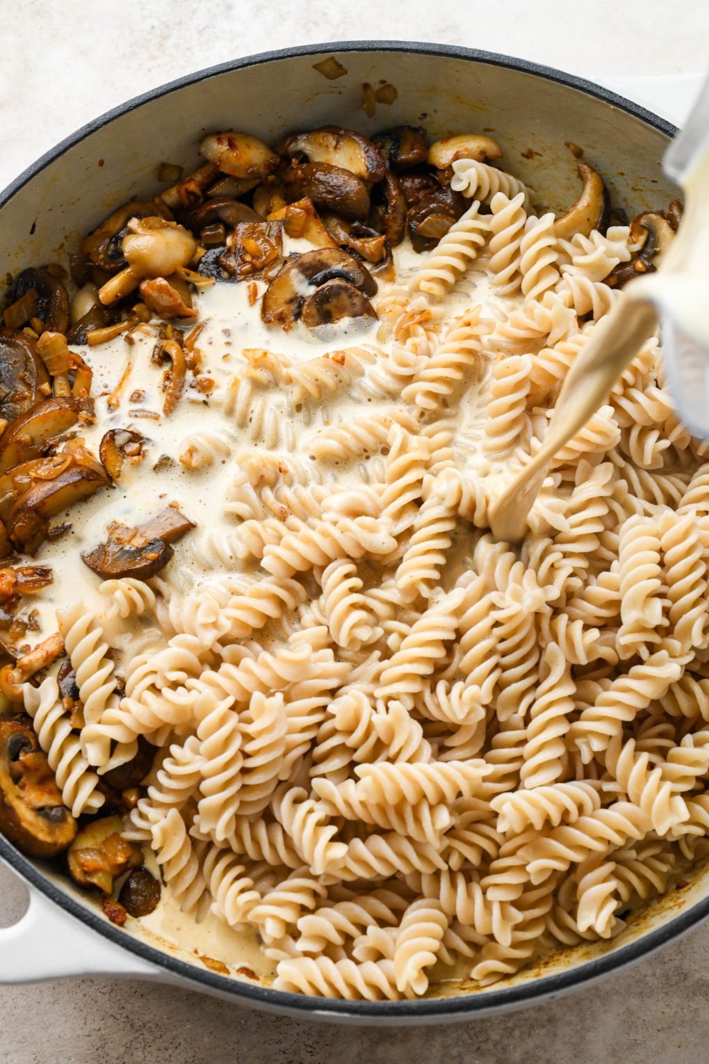 How to make creamy vegan mushroom stroganoff: Pouring cashew cream sauce into a large ceramic skillet with cooked mushrooms and pasta.