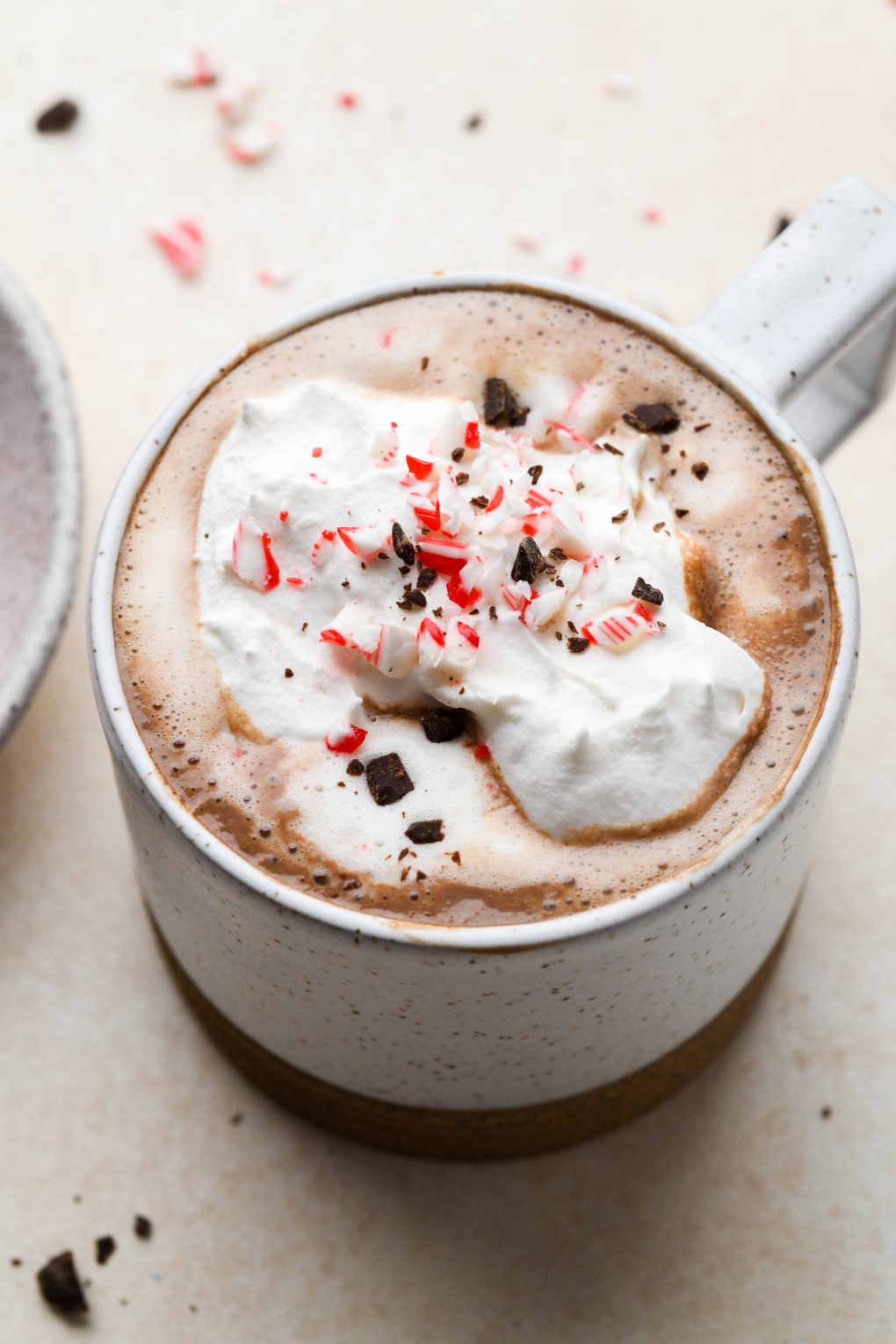 Side view of creamy peppermint hot chocolate topped with whipped cream, crushed candy cane, and shaved chocolate. In a speckled mug on a light brown background. 