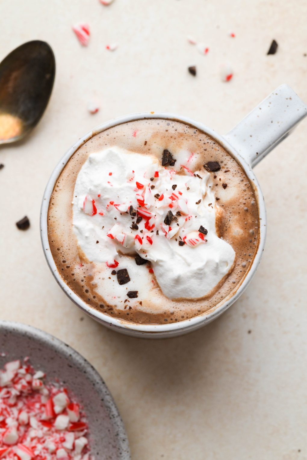 Creamy peppermint hot chocolate topped with whipped cream, crushed candy cane, and shaved chocolate. In a speckled mug on a light brown background. 