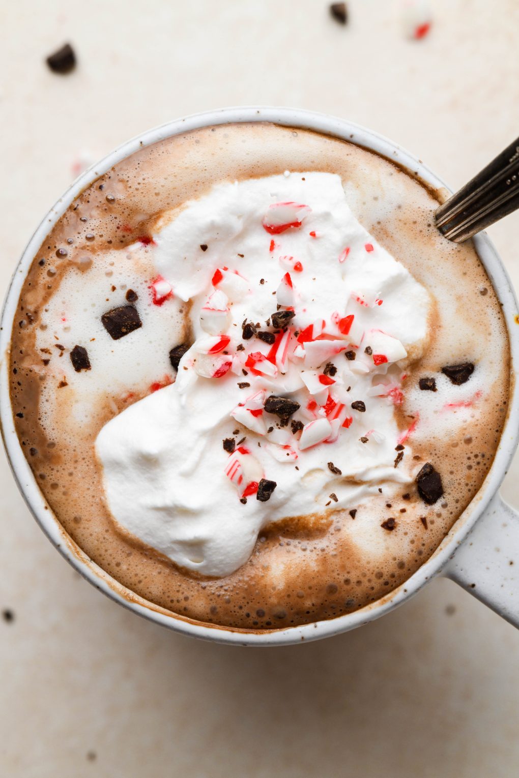Close up shot of creamy peppermint hot chocolate topped with whipped cream, crushed candy cane, and shaved chocolate. In a speckled mug on a light brown background. 