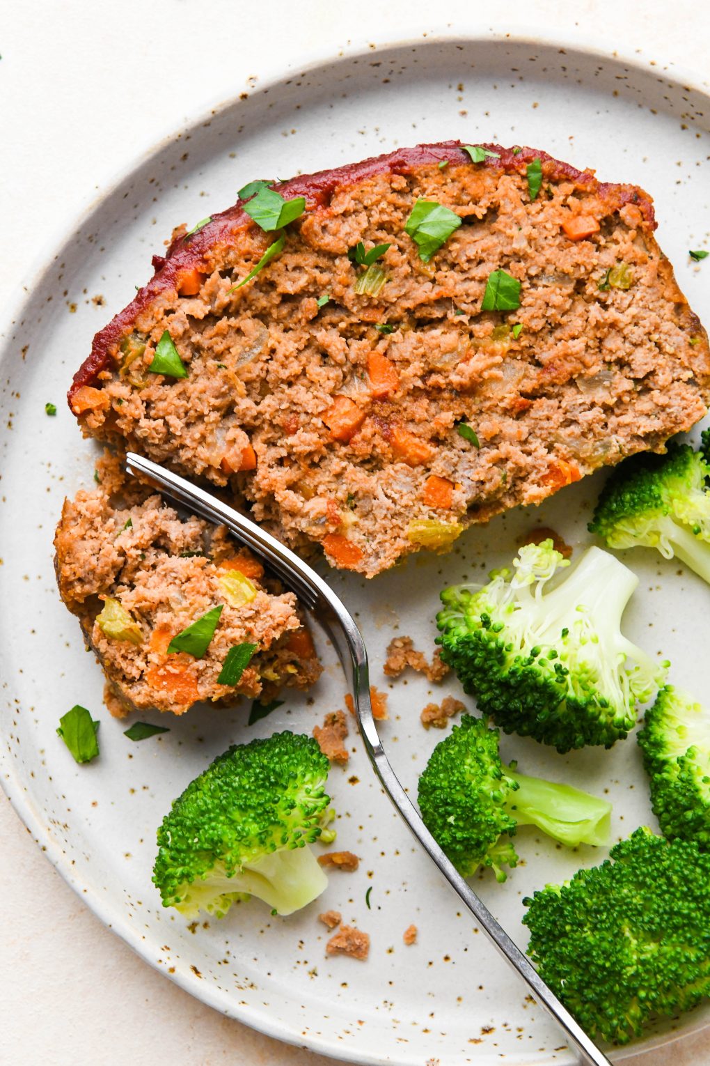 Close up image of a fork cutting into a slice of Whole30 meatloaf on a speckled plate next to a serving of steamed broccoli.