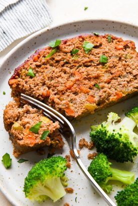 A fork breaking off a bite of Whole30 compliant meatloaf. On a speckled plate next to a serving of steamed broccoli.