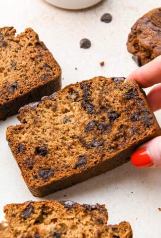 A hand lifting up a slice of easy gluten free banana bread, on a cream colored surface next to several other slices of banana bread.
