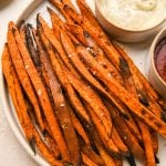 A large speckled plate filled with crispy roasted sweet potato fries next to a ramekin of aioli and a ramekin of ketchup. On a light cream colored surface.