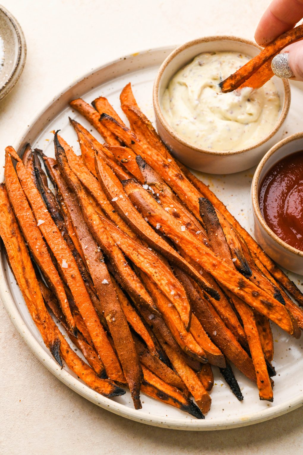 A large speckled plate filled with crispy roasted sweet potato fries with a hand dipping several fries into aioli. On a light cream colored surface.