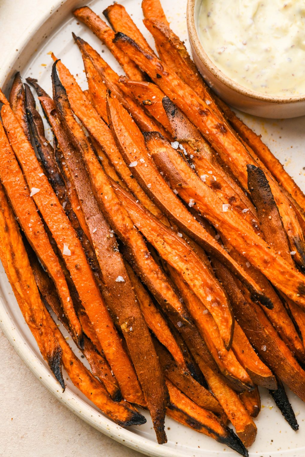 A large speckled plate filled with crispy roasted sweet potato fries next to a ramekin of aioli and a ramekin of ketchup. On a light cream colored surface.