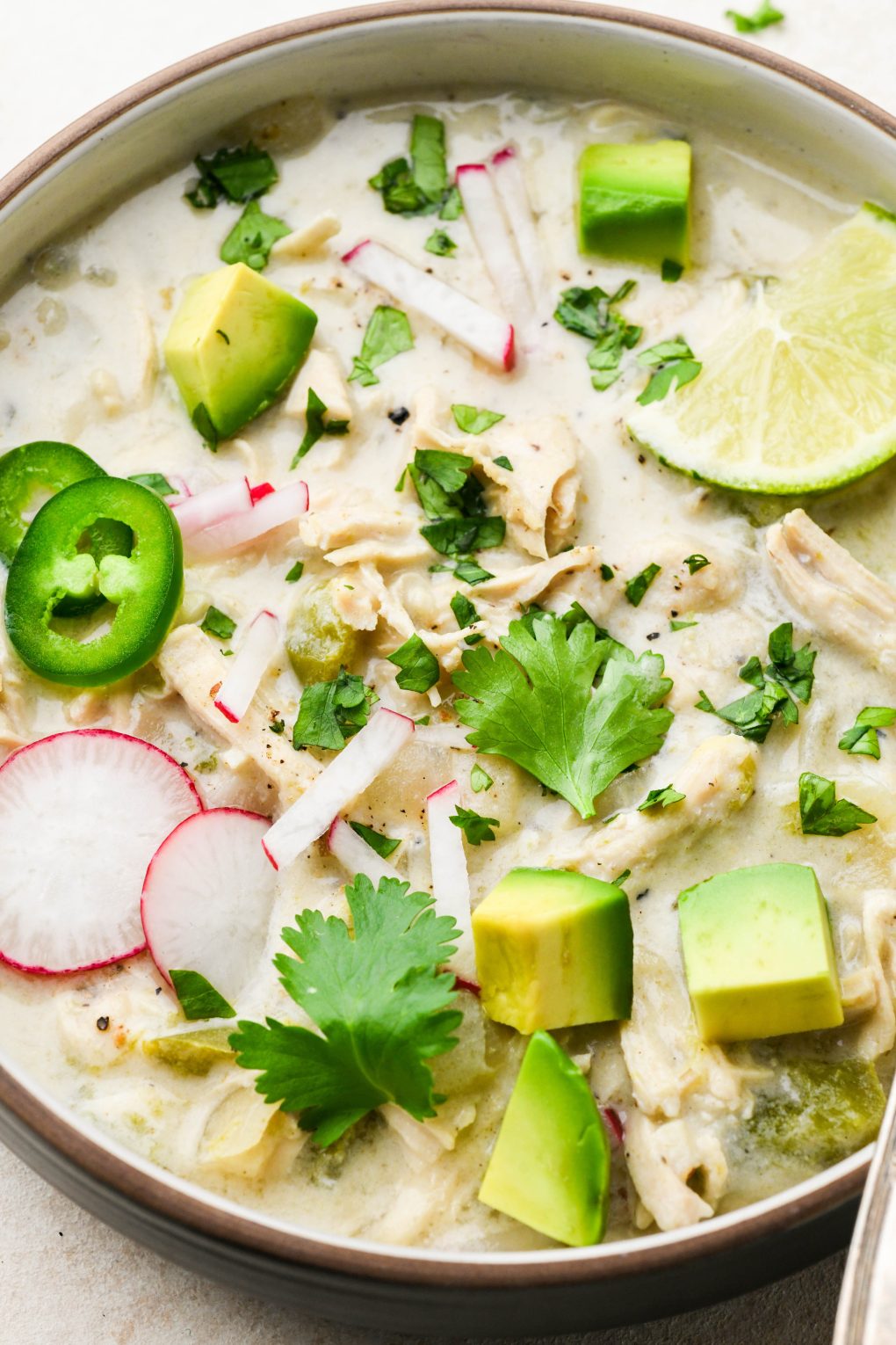 Close up image of a wide ceramic bowl filled with creamy whole30 white chicken chili. Topped with avocado, radishes, jalapeno, cilantro, and lime wedges.
