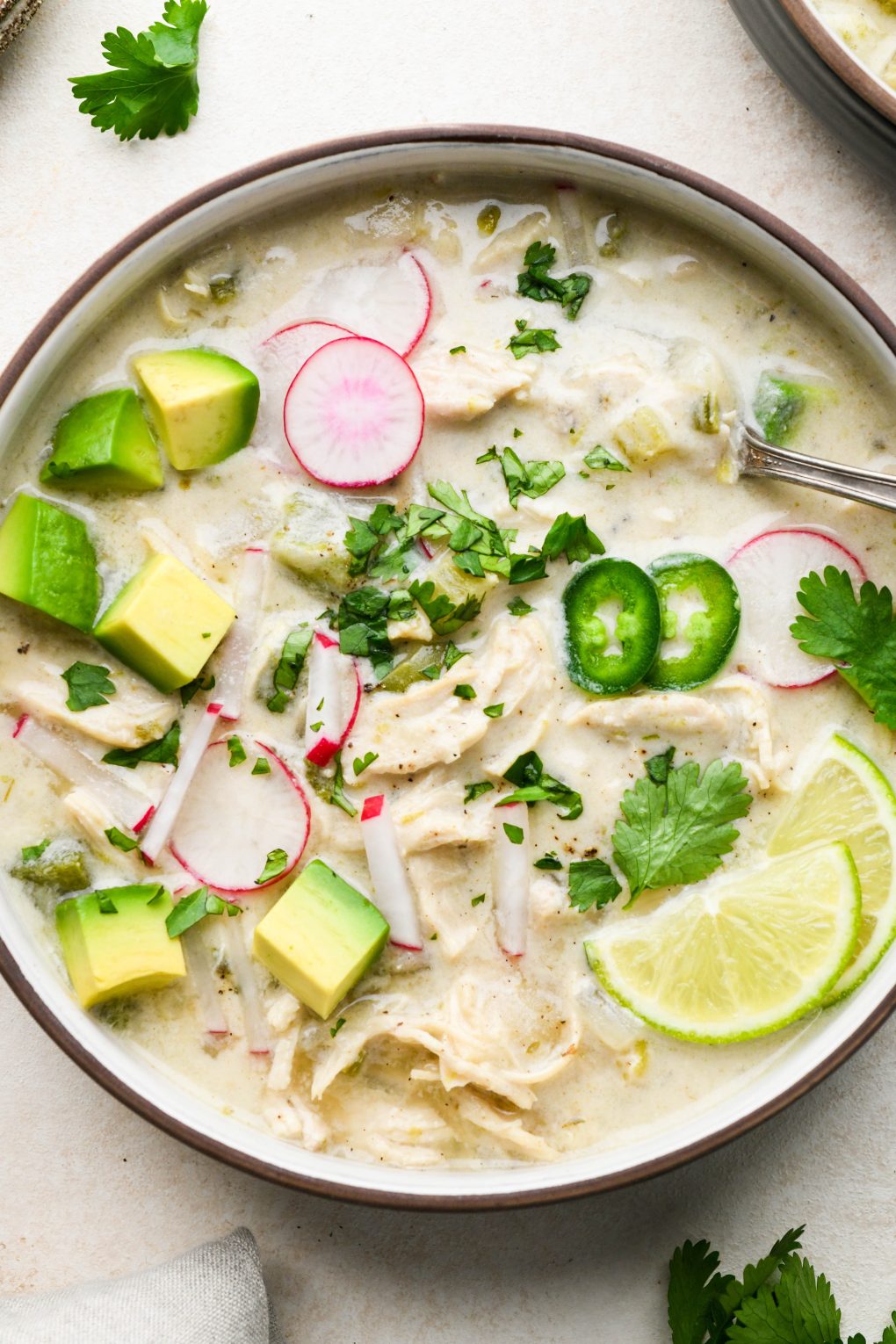 Wide bowl of creamy Whole30 white chicken chili topped with radishes, avocado, cilantro, and lime wedges. On a light cream background surrounded by some scattered herbs. 