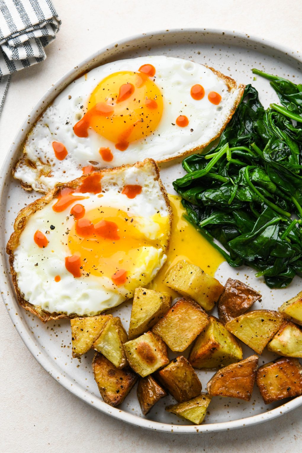 Whole30 breakfast on a speckled plate: Sunny side up fried eggs with hot sauce, sautéed spinach, and roasted potatoes.