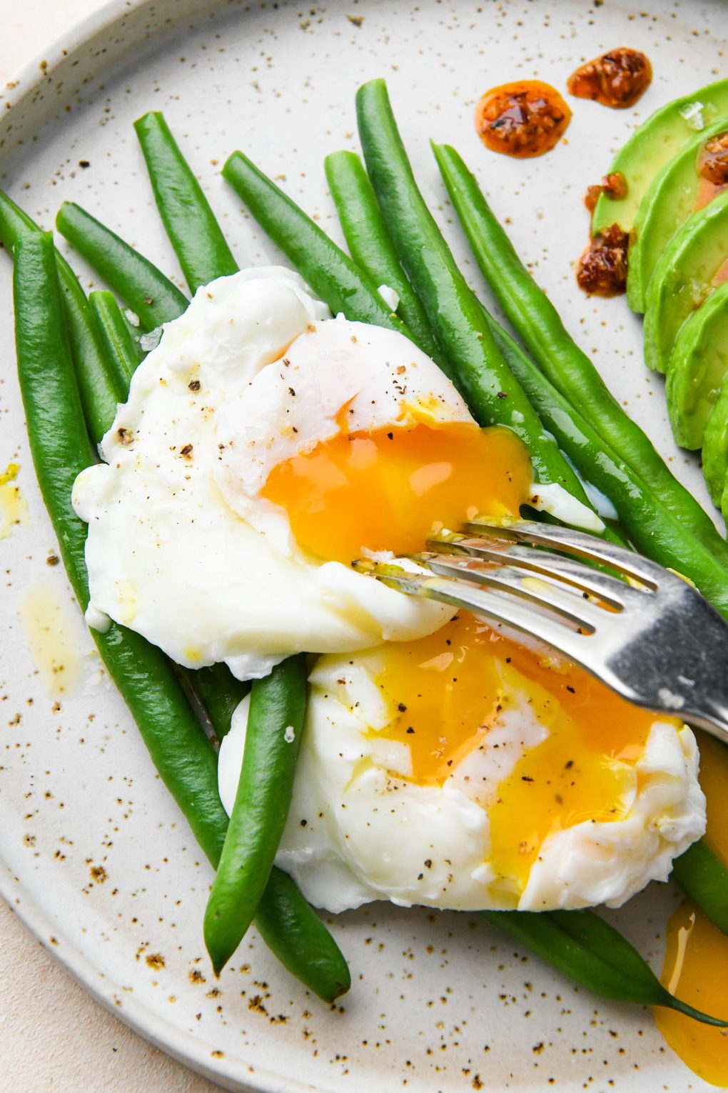 Whole30 breakfast on a speckled plate: Close up of two poached eggs on top of steamed green beans with some thinly sliced avocado on the side. 