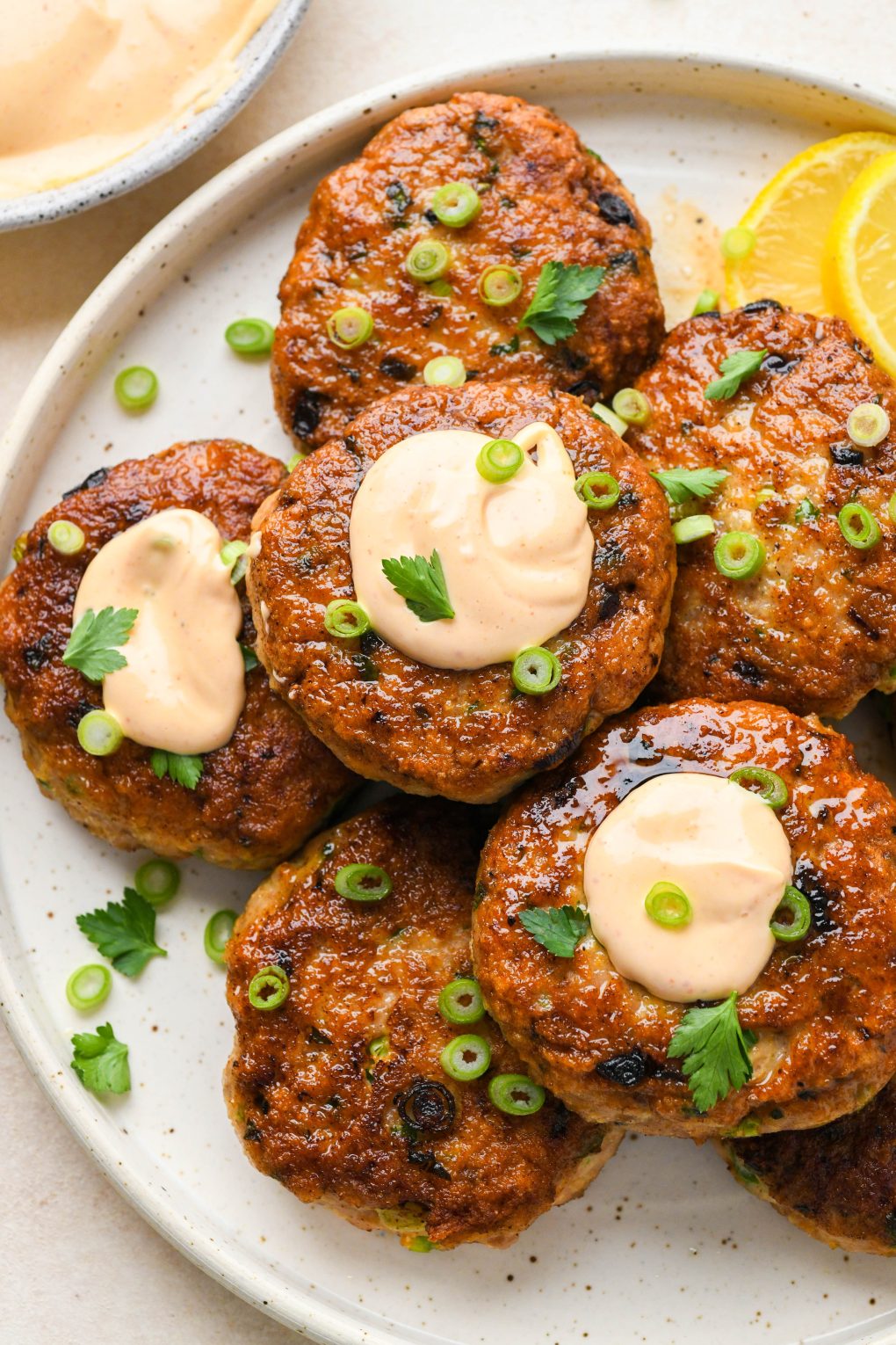 Cooked healthy chicken patties on a speckled ceramic plate topped with dollops of spicy mayo, fresh herbs, and green onions. 