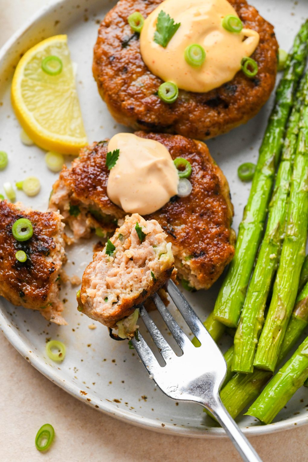 Two cooked healthy chicken patties on a speckled ceramic plate topped with dollops of spicy mayo, fresh herbs, and green onions. One is cut into with a fork showing the juicy interior. Next to a serving of roasted asparagus. 