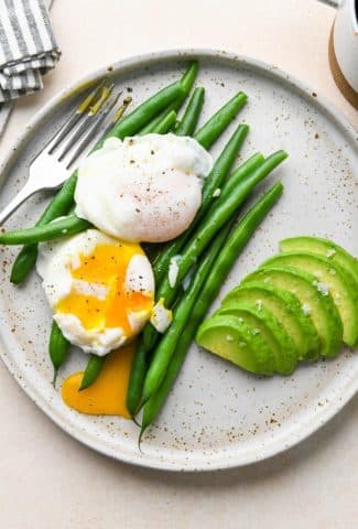 Whole30 breakfast on a speckled plate:Two poached eggs on top of steamed green beans with some thinly sliced avocado on the side.