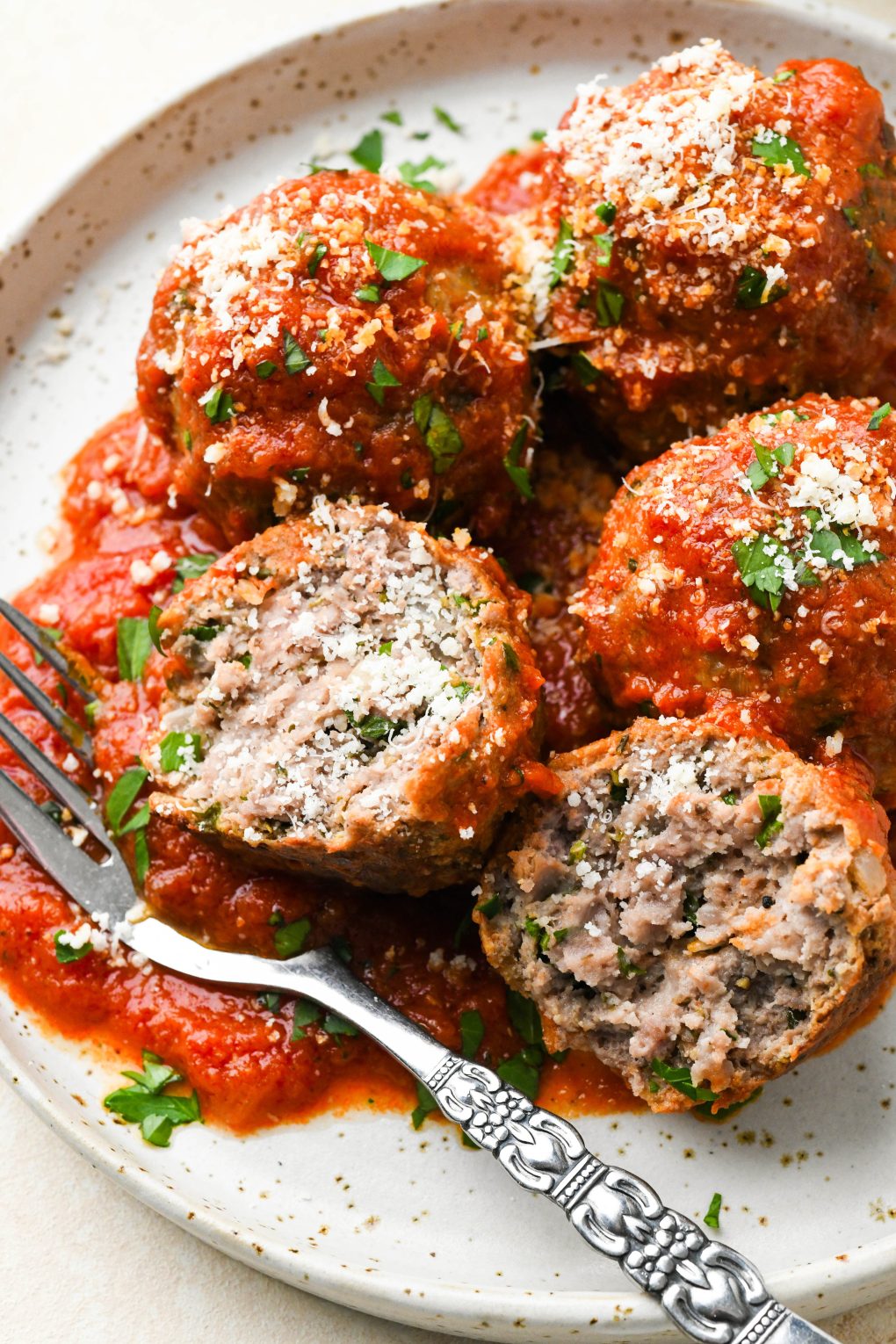 Four gluten free meatballs on a small ceramic plate, one cut open to expose the interior. Topped with grated parmesan and chopped parsley. 