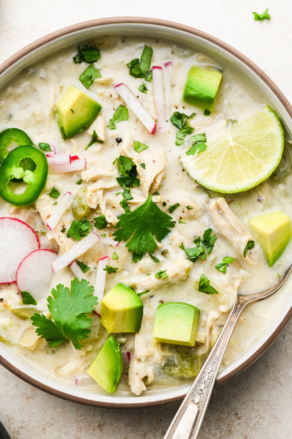 Wide bowl of creamy Whole30 white chicken chili topped with radishes, avocado, cilantro, and lime wedges. On a light cream background.