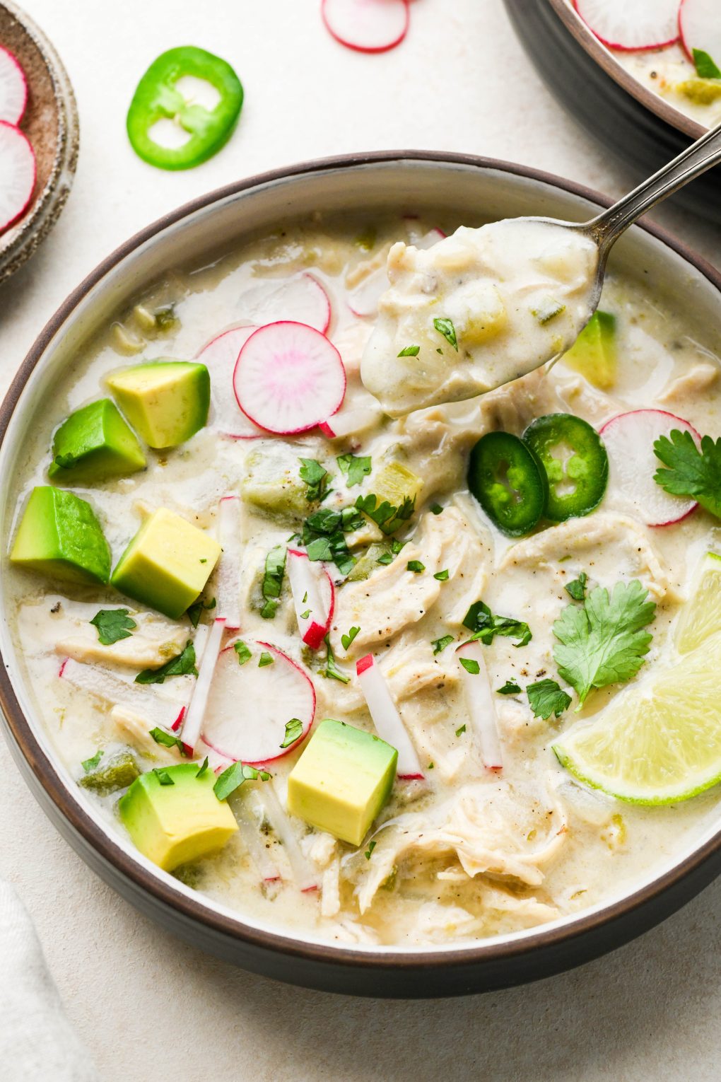 Wide bowl of creamy Whole30 white chicken chili topped with radishes, avocado, cilantro, and lime wedges. A spoon is lifting out one creamy bite. On a light cream background surrounded by some scattered herbs. 