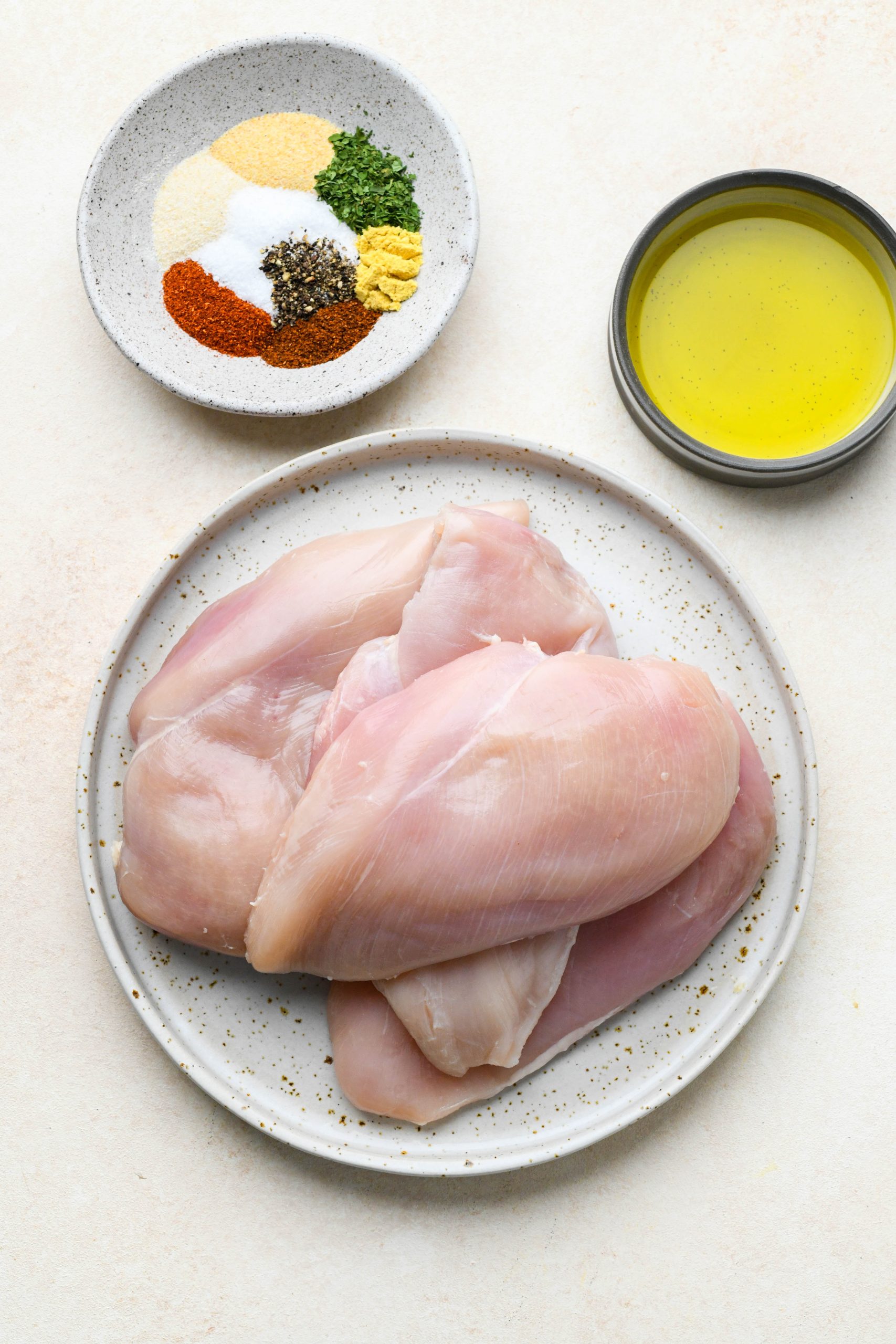 Ingredients for baked chicken breast recipe on various ceramics, on a light cream colored background.