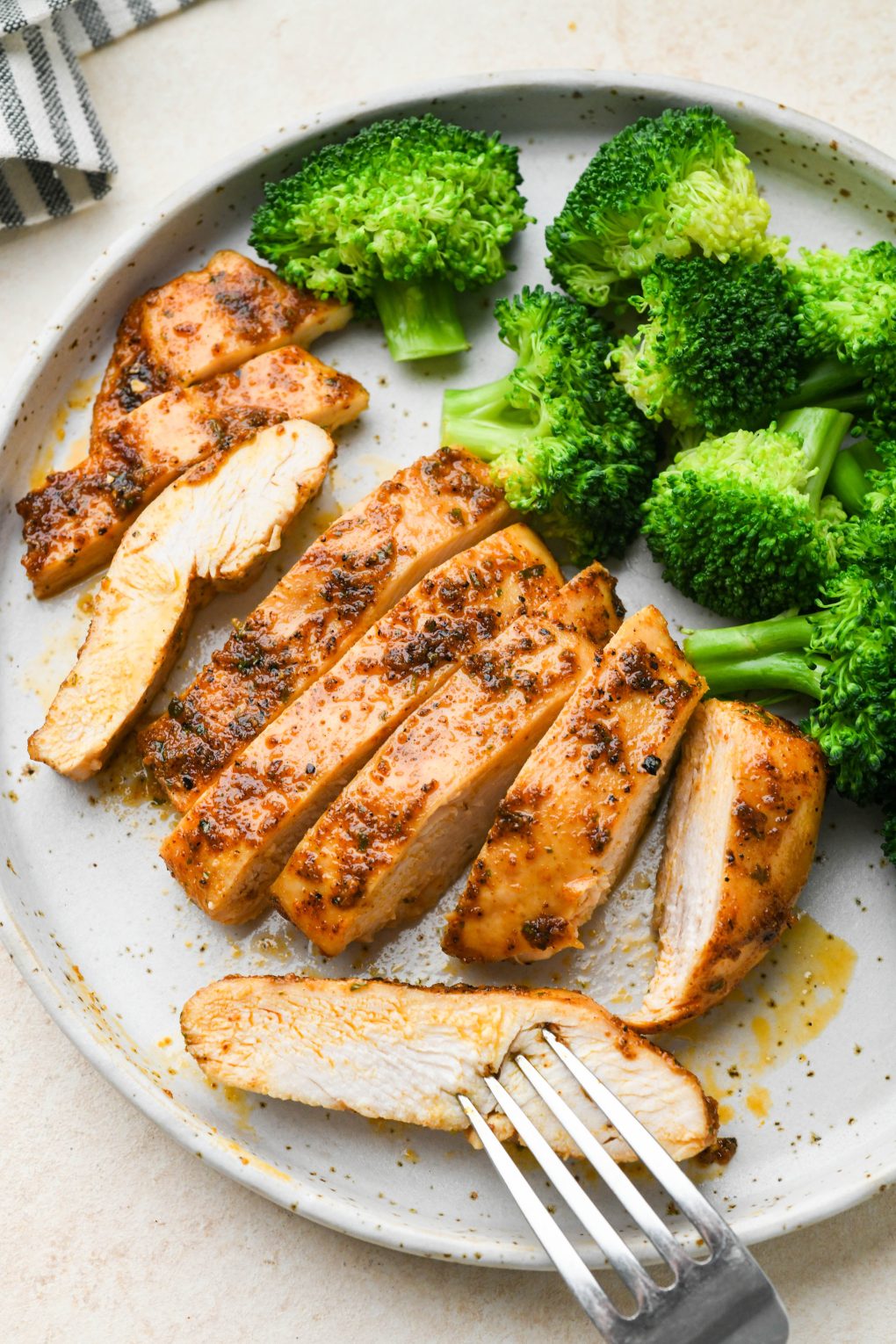 Baked chicken breast sliced on a light colored speckled plate, next to some steamed broccoli.