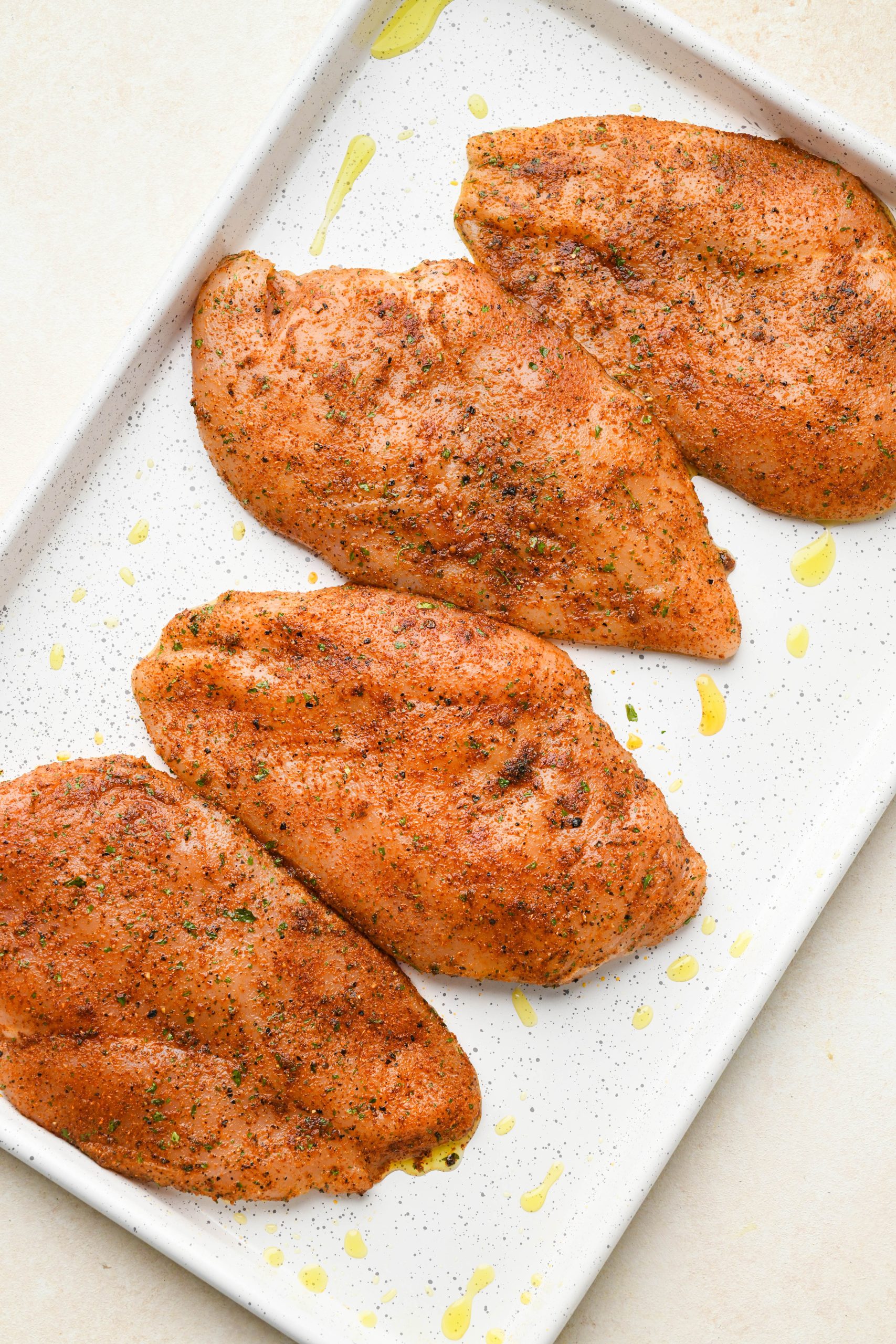 How to make baked chicken breast: Raw spice coated chicken breasts evenly spaced out on a large rimmed baking sheet.