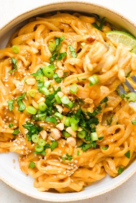 Red curry peanut noodles piled into a small ceramic bowl and topped with cilantro, green onions, sesame seeds, and chopped peanuts. A fork is lifting out a bite from the tangle of noodles.