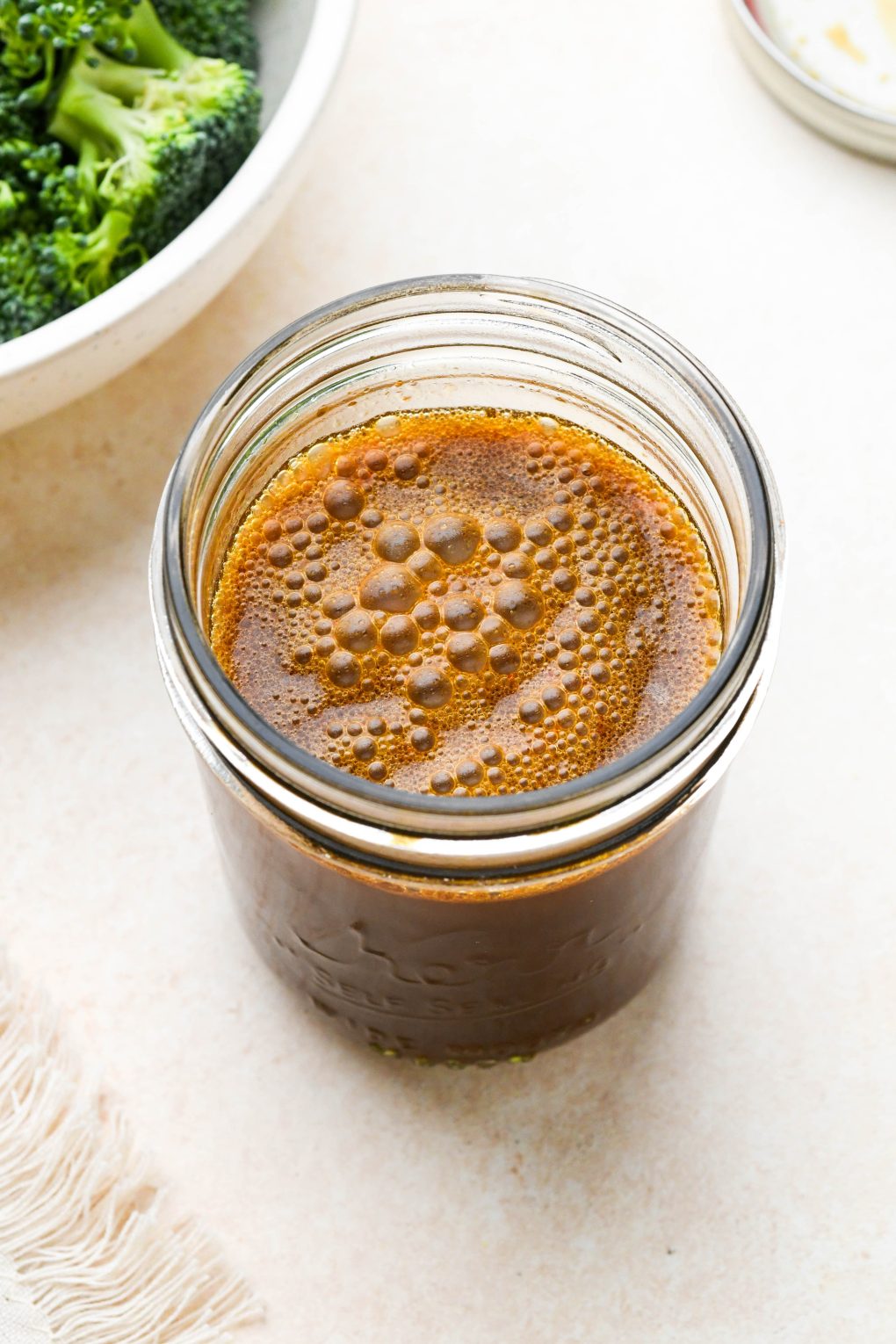 A small glass ball jar of healthy homemade stir fry sauce, next to a bowl of broccoli florets.

