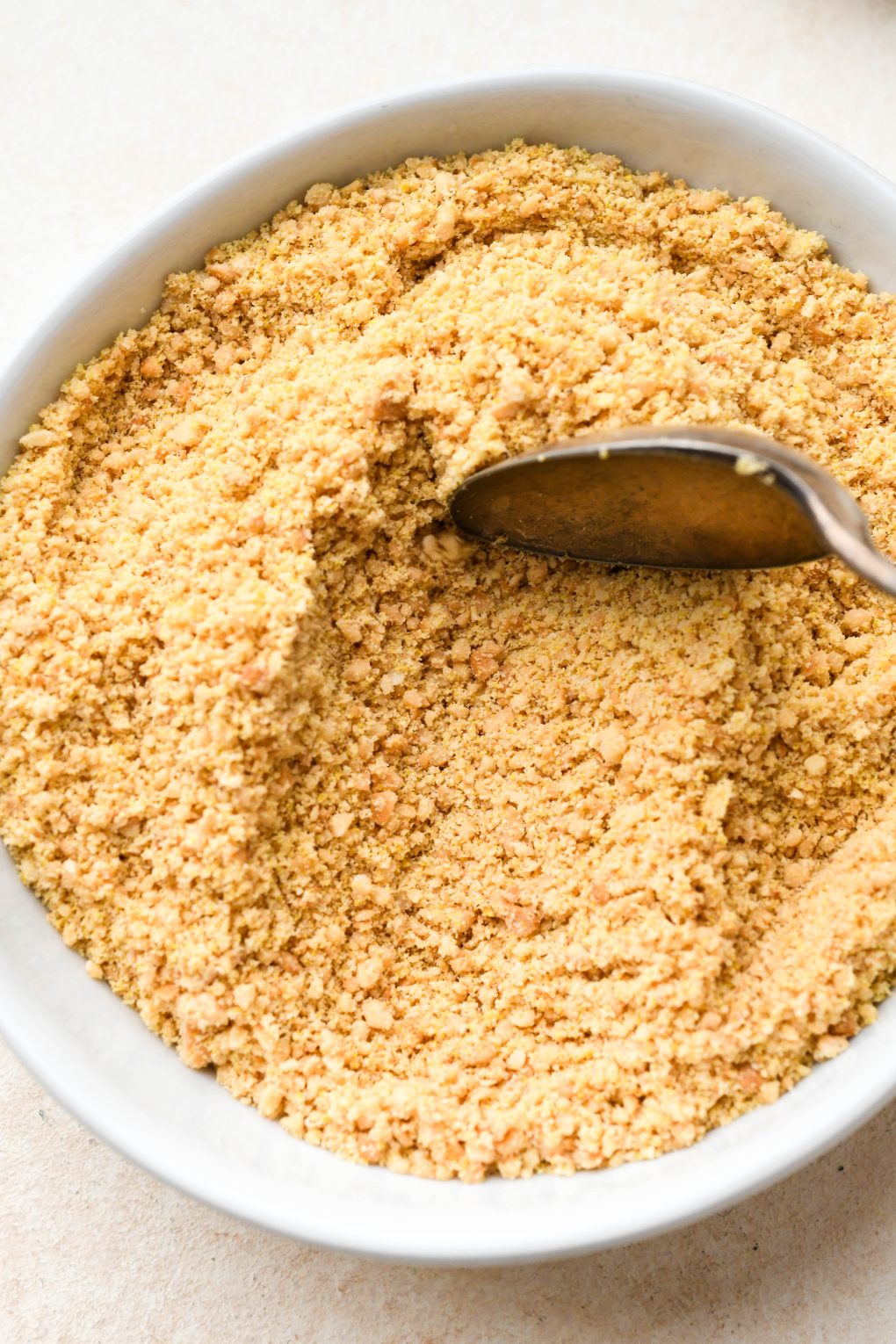 Vegan parmesan cheese in a small shallow white bowl with a spoon dragging through it to show the texture, on a light cream colored background.
