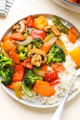 Colorful veggie stir fry on a wide shallow ceramic plate with white rice, topped with sesame seeds.