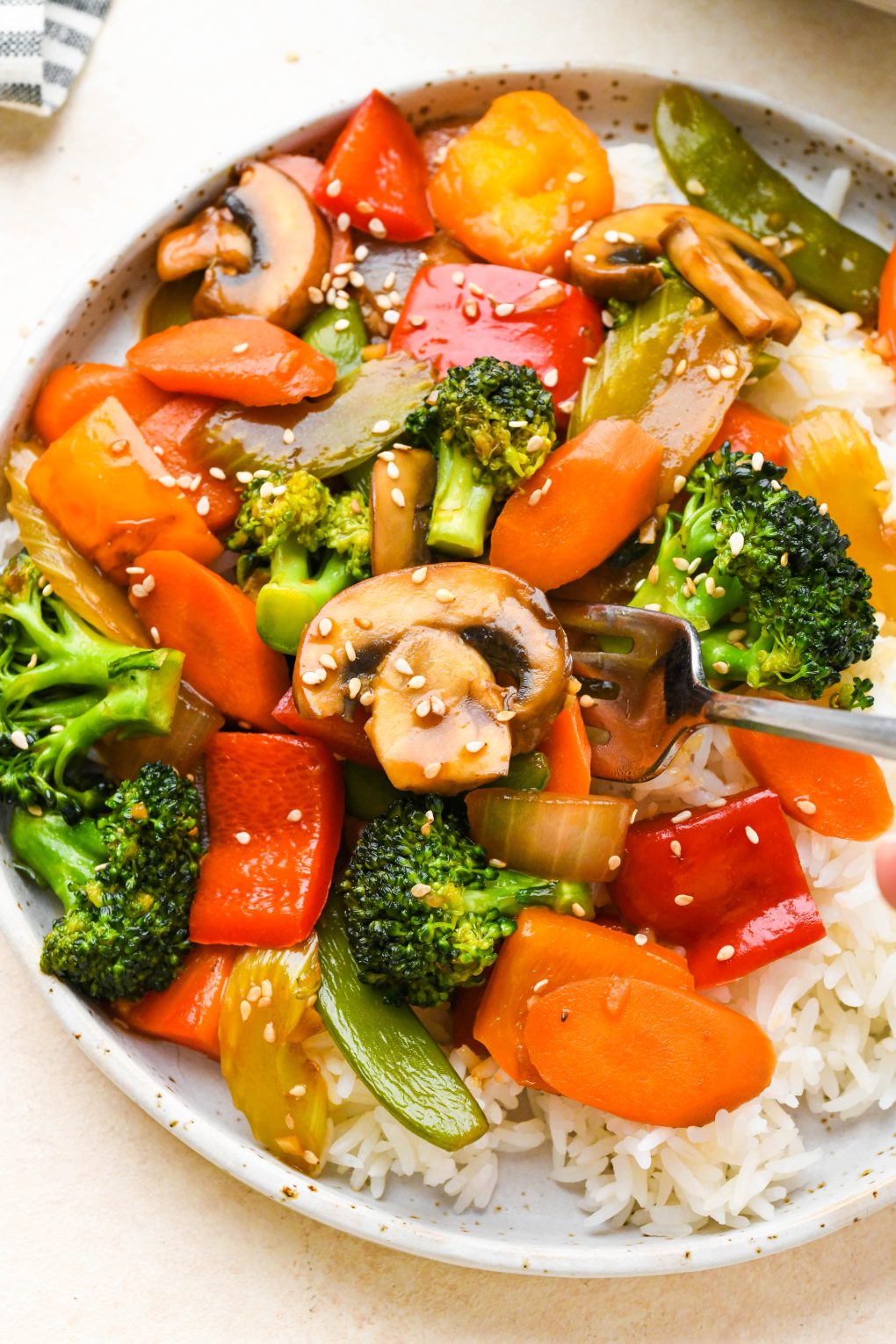 Close up of a colorful veggie stir fry on a wide shallow ceramic plate with white rice, topped with sesame seeds.