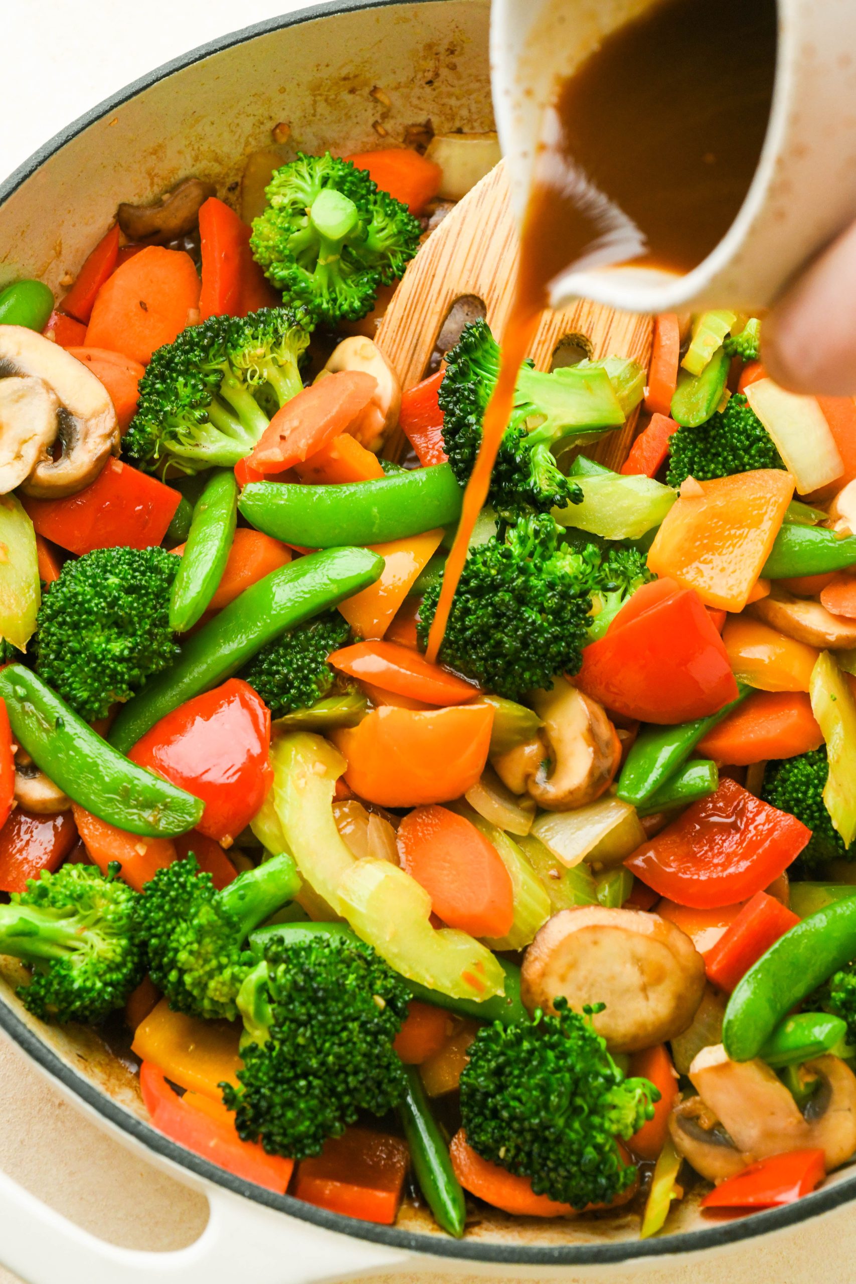 How to make easy veggie stir fry: Slightly cooked vegetables in a large skillet with a hand pouring in stir fry sauce.