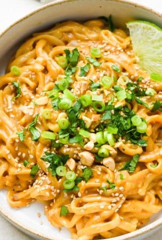 Red curry peanut noodles piled into a small ceramic bowl and topped with cilantro, green onions, sesame seeds, and chopped peanuts.