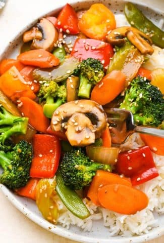 Close up of a colorful veggie stir fry on a wide shallow ceramic plate with white rice, topped with sesame seeds.