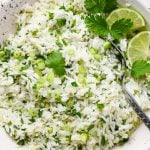A large speckled ceramic bowl of cilantro lime rice, topped with thinly sliced green onion, cilantro leaves, and lime wedges.