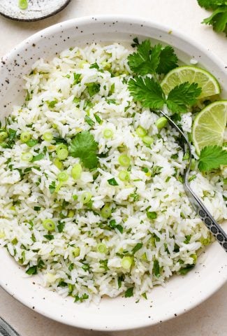 A large speckled ceramic bowl of cilantro lime rice, topped with thinly sliced green onion, cilantro leaves, and lime wedges.