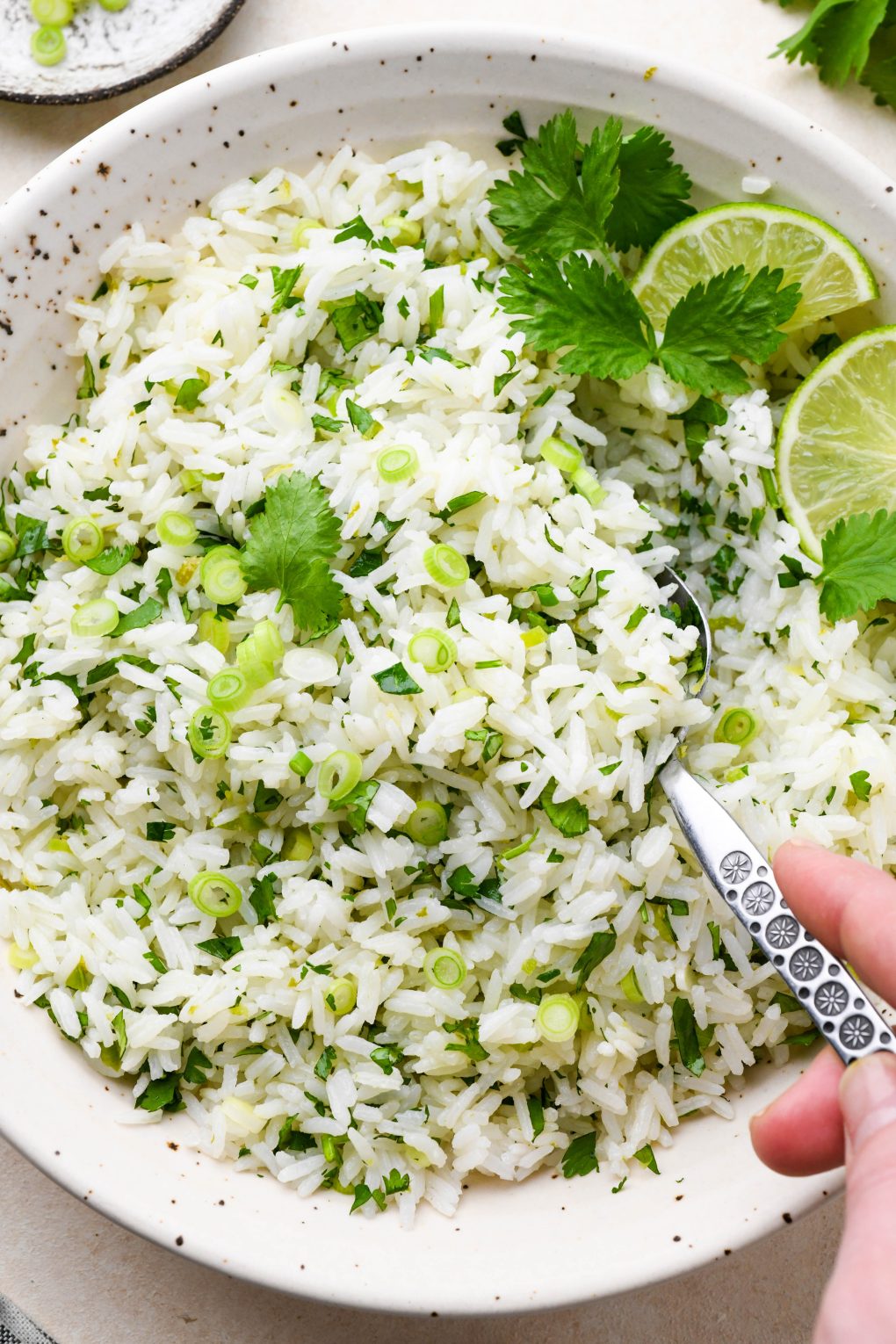 A large speckled ceramic bowl of cilantro lime rice, topped with thinly sliced green onion, cilantro leaves, and lime wedges, with a spoon lifting some rice out of the bowl.