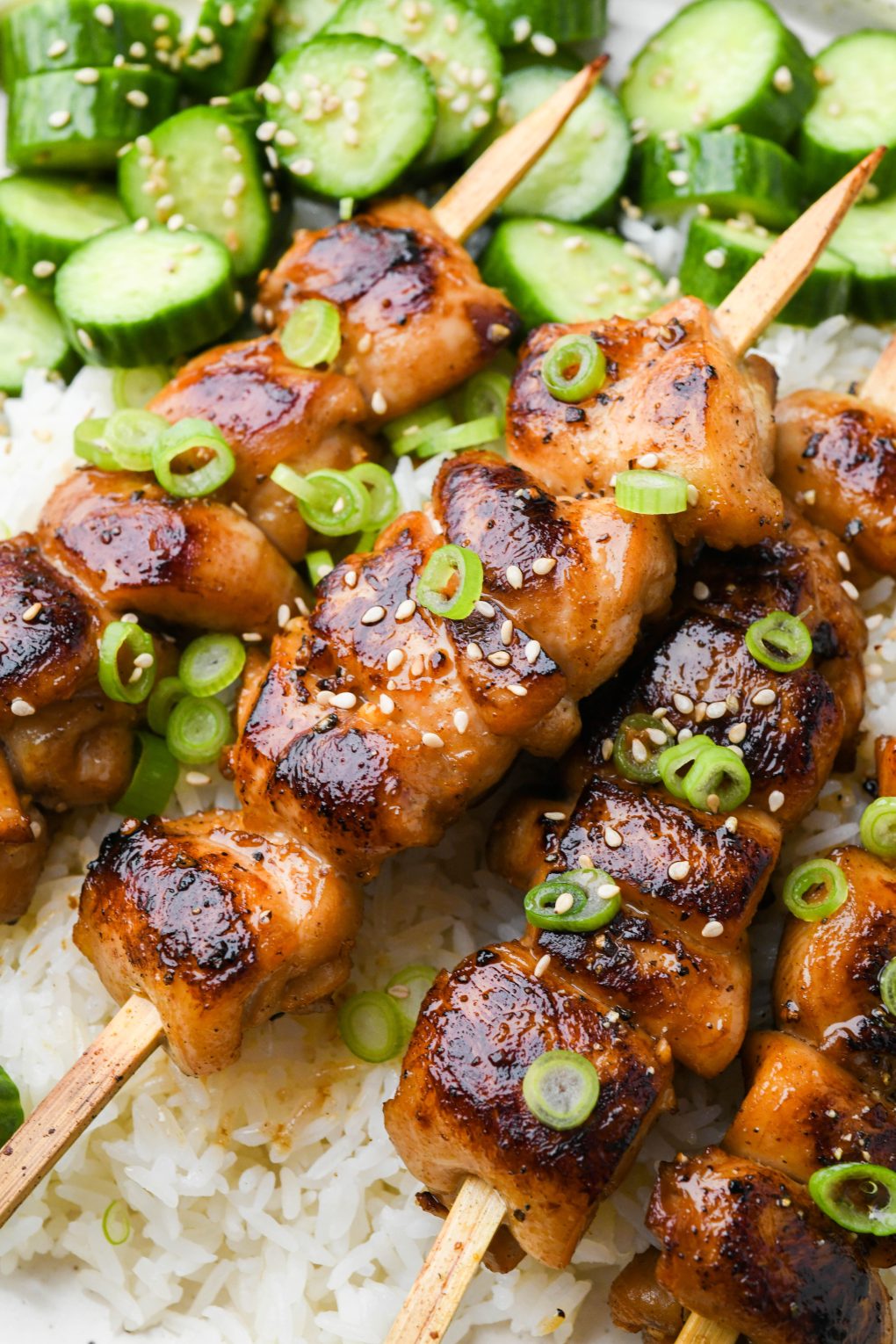 Close up of grilled garlic ginger chicken skewers on top of white rice, next to an asian cucumber salad.