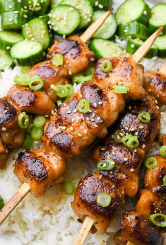 Close up of grilled garlic ginger chicken skewers on top of white rice, next to an asian cucumber salad.
