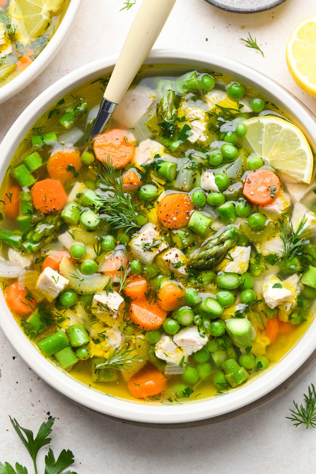 A shallow ceramic bowl filled with chicken soup with spring vegetables - peas, carrots, celery, and asparagus. Garnished with fresh dill and lemon wedges.