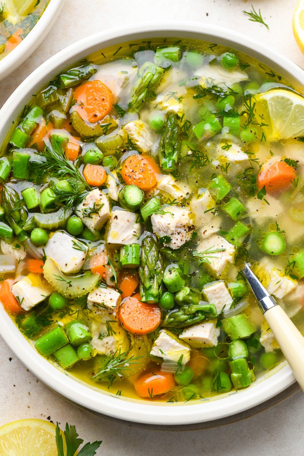 A shallow ceramic bowl filled with chicken soup with spring vegetables - peas, carrots, celery, and asparagus, with a spoon dipping in. Garnished with fresh dill and lemon wedges.