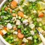 A shallow ceramic bowl filled with chicken soup with spring vegetables - peas, carrots, celery, and asparagus, with a spoon dipping in. Garnished with fresh dill and lemon wedges.