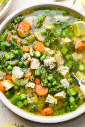 A close up image of a shallow ceramic bowl filled with chicken soup with spring vegetables - peas, carrots, celery, and asparagus. Garnished with fresh dill and lemon wedges.