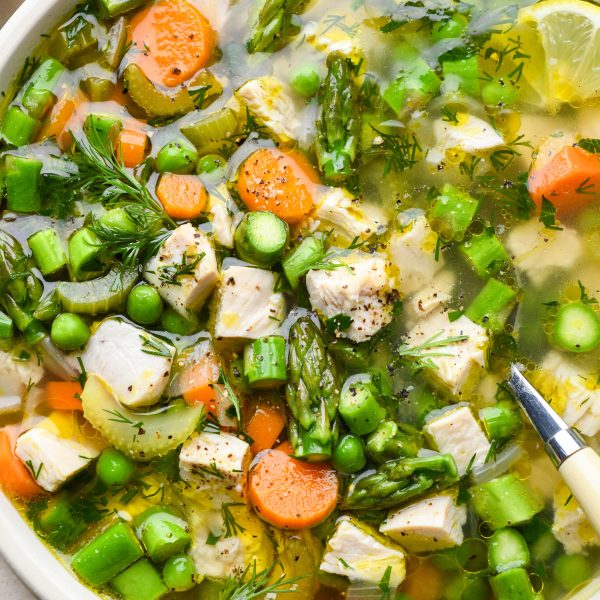 A close up image of a shallow ceramic bowl filled with chicken soup with spring vegetables - peas, carrots, celery, and asparagus. Garnished with fresh dill and lemon wedges.