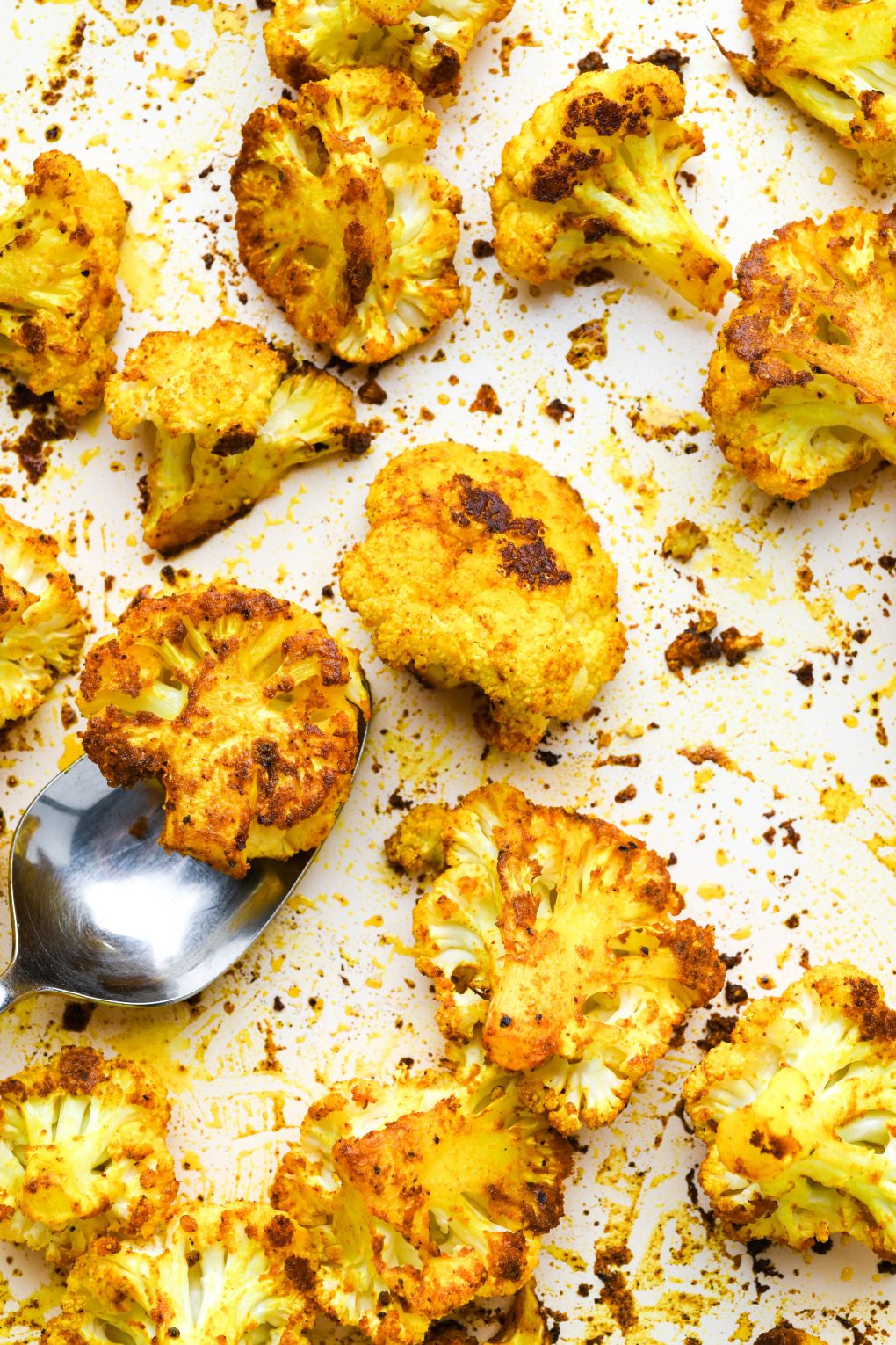 Turmeric roasted cauliflower on a white baking sheet with a metal spoon scooping up a single piece of caramelized cauliflower.
