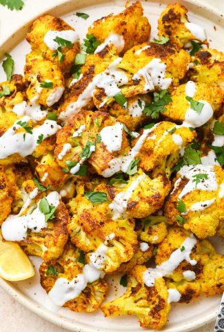 Turmeric roasted cauliflower florets on a speckled plate topped with a drizzle of thinned yogurt and fresh herbs.